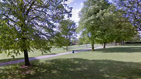 Google A large park in sunshine with trees in leaf and a bench and bin alongside a pathwway