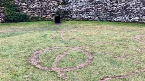 A number of tyre tracks are seen on grass. The grass is inside a stone fort, the walls of which are viewed in the background.