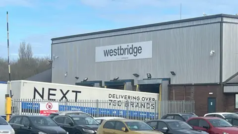 A factory building with a sign saying 'westbridge'. A next lorry is parked outside. Multiple cars are parked in a fenced off car park next to it. 