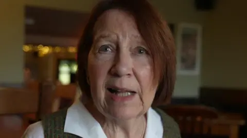 Heather Swettenham is sat in the George & Dragon pub in Hudswell. She is wearing a white shirt with a green jumper over it.