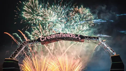 The Creamfields logo at the entrance of the festival site is displayed in front of the night sky lit up with yellow, pink and green fireworks
