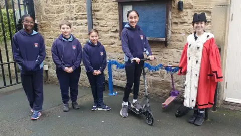 Five children are standing outside in front of a stone wall with a blue noticeboard, four of them wearing matching navy school hoodies. One child is dressed in a bright red ceremonial robe and black hat, and two scooters are parked beside them.