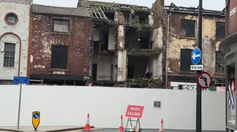 Michael Broomhead/LDRS A row of derelict buildings which are cordoned off by a white wall. The interior of one building is visible as the frontage has fallen away. There is a sign on the road which says 'Road closed' and three orange and white cones.