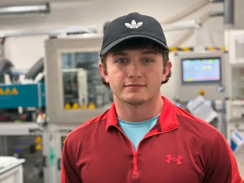 BBC/Richard Moss Jack Mason-Young looks at the camera. He has brown hair and is wearing a black baseball cap and red sweater. Behind him is lab machinery.
