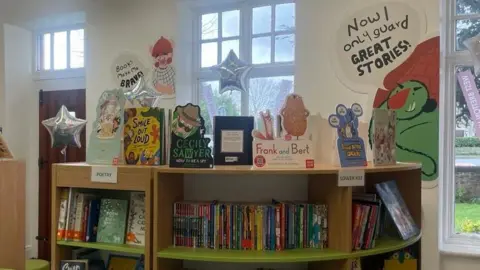 Sofía Luis-Hobbs/BBC Library shelves full of colourful books in front of a white wall that has drawings of book characters. One green character has a speech bubble appearing from its mouth that reads 'now I only guard great stories!'. To its left, a cartoon pig on the wall has a speech bubble that reads 'books make me brave!'