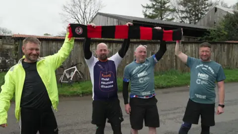 Four men holding up a Truro United scarf as they arrive in Truro, Cornwall. The scarf is red and black. They are all smiling at the camera. 