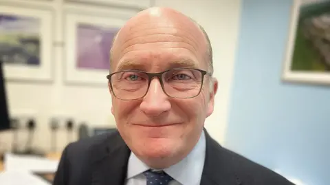 Nikki Fox/BBC Nick Hulme smiles at the camera. He is bald and wears glasses, a black suit and white shirt with a tie.