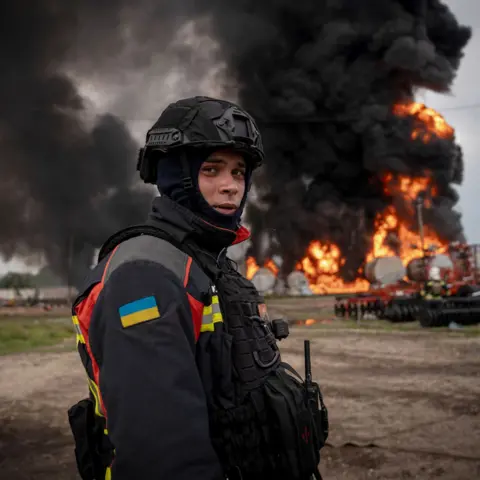 Valeria Demenko/DSNS A member of the emergency services in north-eastern Sumy, dressed in high vis with the Ukraine flag sewn onto his arm and a combat helmet and balaclava, looks wearily at the camera while standing in front of a burning gas depot near the Russian border.