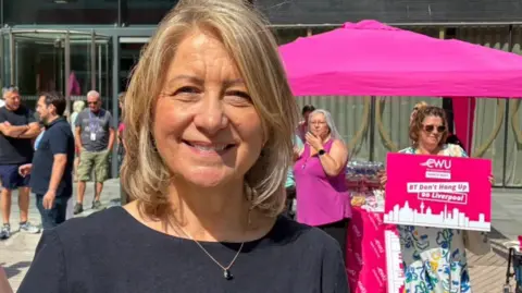 Karen Rose with bobbed blond hair wears a black t-shirt. She stands outside the Plaza building in Liverpool city centre with protesters with a hot pink marquee standing behind her.