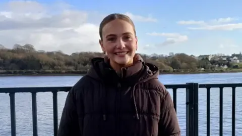 A woman smiling at the camera, she is wearing a brown coat. She has dark hair.