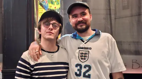 George Wright, wearing a Euro 96 England shirt with 32 on the front, puts his arm around his brother, Laurie, wearing a stripey long-sleeve t-shirt while standing outside a pub in central London. 