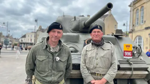Two men in khaki jackets and berets stand in front of a tank, as part of VE day anniversary celebrations. Both are smiling at the camera. 