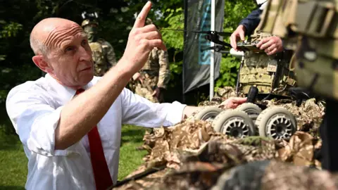 efence Secretary John Healey gestures to the drone in the sky as he views a screen showing it’s camera feed