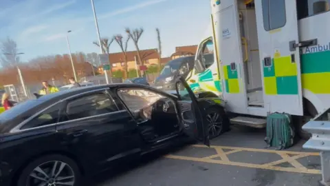 BBC A black car shown with its door open against the side of an ambulance