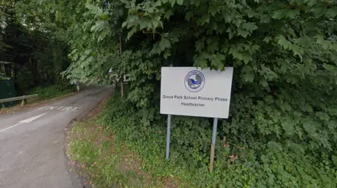 A white sign with the Grove Park School logo, a blue bird and grey trees in a blue circle, and text that reads Grove Park School Primary Phase, followed by Headteacher. The sign is situated in front of leaves that fill most of the frame and a tarmac road marked as entry with paint.