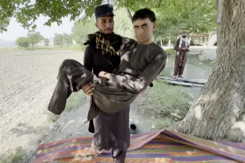 BBC One of the 12 migrants who spoke to BBC Afghan Forensic, quadruple amputee Shahsawar, 21, is carried by another man, on a red-and-blue striped rug, while a third looks on, in a tree-lined Afghan landscape. All wear brown salwar-kameez, the two unnamed, bearded, men hats and patterned scarves.