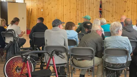 In the foreground is a wheel chair and mobility walker - in the centre of the picture are Karen John and Wayne facing away, sat in fold up chairs. The chairs are lined up facing the front with lots of other families waiting for someone to talk