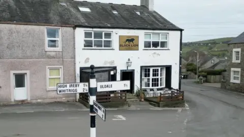 BBC Corner view of a pub - the Black Lion. It is a white building, with a bay window to the right of the front door. On either side of the door is a small, fenced off, raised area with chairs and tables. The building is located on a street corner, with a pebble dashed property to the left, and the street leading down from the right offering a view of hills to the side. A sign post measuring distances to High Ireby, Whitrigg and Uldale is at the front.