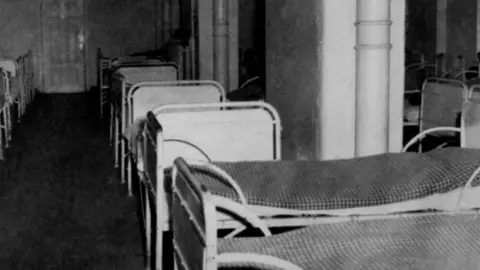 The reception room of the psychiatric clinic at Hadamar where those detained waited for mortal injections as part of Aktion T4. Several single beds can be seen lined up beside each other in a black and white image.