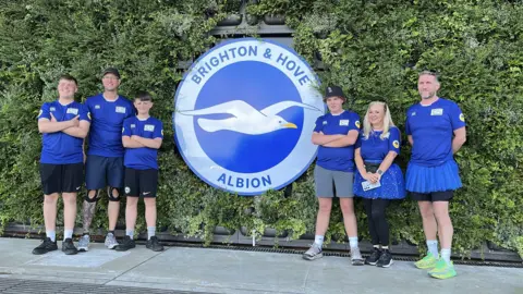 George Carden/BBC Six people stood in front of a hedge, which has a huge sign bearing Brighton and Hove Albion's blue and white seagull logo. The people, three of whom are teenagers and three others are older, all wear matching blue t-shirts and the man and woman to the right of the image are wearing blue tutu skirts.