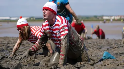 Sonia Watson and Ben Fryer crawling through deep mud while wearing Where's Wally outfits, with red and white striped tops and hats.