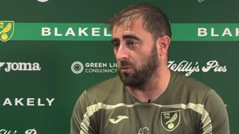 Andy Cook in a green football shirt speaking at a press conference.