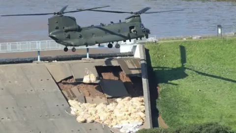 PA Media A chinook helicopter dropping beige bags on the side of a dam
