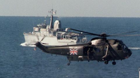 A Royal Navy Sea King helicopter hovers over the sea with a warship in the background