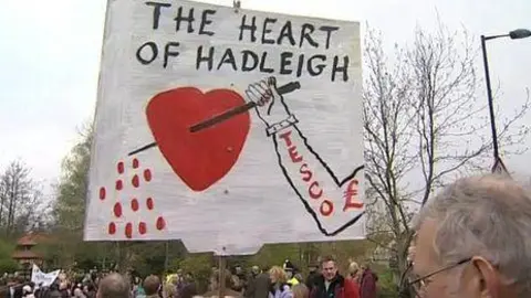 A sign is held up in a crowd that shows the words "The heart of Hadleigh" with an image of an arm holding a spike piercing a heart, Tesco is written on the arm with a pound symbol next to it.