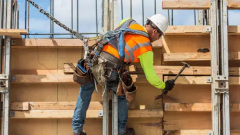 US construction worker using a hammer on a building site. He is tethered to the side of the building and is wearing a tool belt, hi-vis clothing and a hard hat.