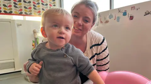 Baby Lennon is 13 months old and is almost walking. He is held from behind by his mum Ellie. He is wearing a grey sweatshirt. She is wearing a white and black striped cardigan. 