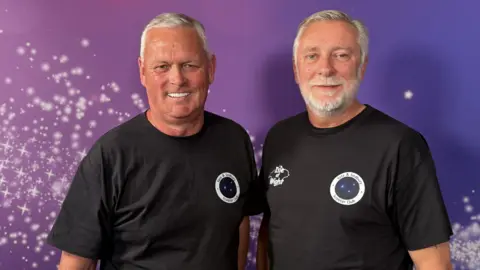 Kevin Milford (left) and Andrew Davis (right) are smiling at the camera. They are both wearing black T-Shirts with the Yate & Chipping Sodbury scooter club logo over their left chest. Kevin has grey, short hair and Andrew has grey, short hair and a grey beard. They are stood in front of a purple backdrop. 