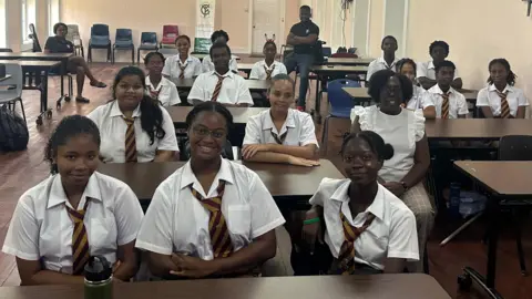 Teenage girls and boys sit behind desks in a classroom at Harrison College. All the pupils are wearing white shirts or blouses and blue-and-yellow striped ties. Two teaching staff, a man and a woman dressed in blue, are at the back of the classroom.