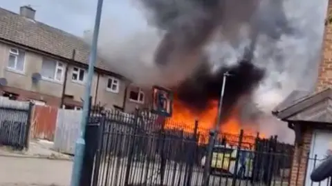 Smoke and fire billow from a end of terrace house, shown behind black metal gates