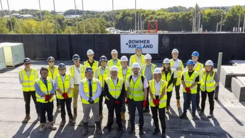 A group of 20 men and woman are standing on the roof space of an apartment block, hearing white and blue helmets and yellow high-visibility jackets. There is a sign in the background that reads "Bowmer and Kirkland".