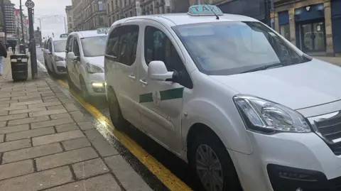 A row of three white taxis lined up at a taxi rank in a city centre street, with shops and street furniture visible in the background