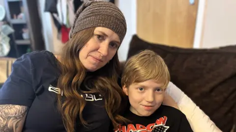 Cheryl and her son Ashton sat on the sofa. Cheryl is wearing a navy t-shirt and is wearing a grey / brown hat. She has tattoos on one arm and she has long brown hair