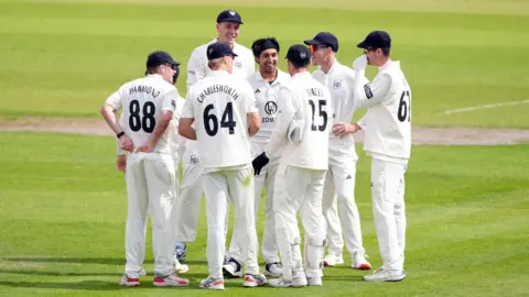 PA Media A photo showing a group of men dressed in cricket whites and wearing hats standing on a green cricket pitch
