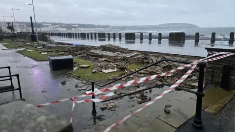 Picture of the damaged Sunken Gardens. Red and white tape marking off the sight can be seen whilst a bricked wall lies on the ground after being blown over. Picture taken on a grey, wet day.