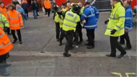 A cluster of police officers in hi-viz and stewards in blue on the concourse of a football ground, surrounding a barely visible football fan