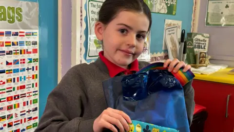 Erin holds her bag, made with blue fabric, a rainbow strap and a small blue and yellow patterned pocket on the front. She is smiling at the camera as she sits in front of a blue classroom wall display.