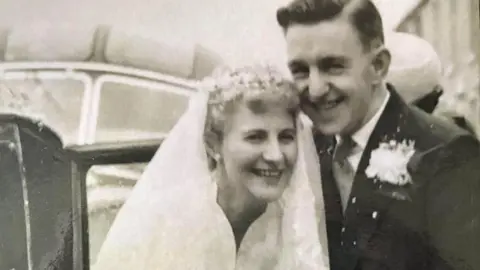 HC-One handout Gordon and Joan Cilgram on their wedding day in 1958 smiling standing in front of a wedding car. Joan with short blond hair wears a white veil and wedding dress and Gordon wears a black suit with a boutonnière 