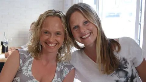 Two women sit together and smile at the camera. Their heads are leaning together and they are smiling. They are in a restaurant and behind them is a table with a wine bottle and wine glass on it.