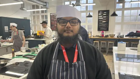BBC A young man in a chef's uniform looks into the camera in a busy training kitchen. He wears a white chef hat, a red lanyard, and a blue and white striped apron. 