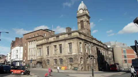 A Victorian stone built building with a clock tower and tall windows covers the corner of a street.
