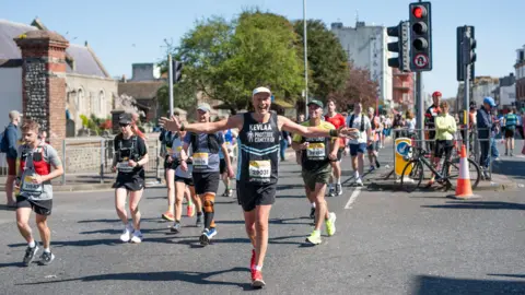 Kevin Webber People are running along the road with one runner, Kevin Webber, in the centre, wearing a black “Prostate Cancer UK” vest and race number 2903. He is running with both arms stretched wide, wearing a white cap and red running shoes. The road is lined with metal barriers and there are spectators in the background.