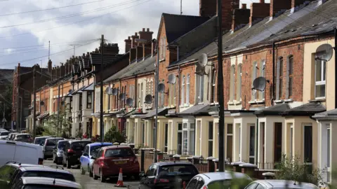 Getty Images A row of terraced houses. Cars are parked outside in the road. Satellite dishes are up outside houses. A cone is sitting on the road. 