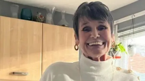 Catherine has short grey hair and is wearing a pink and white jumper. She is standing in front of cabinets in a kitchen and is smiling at the camera.