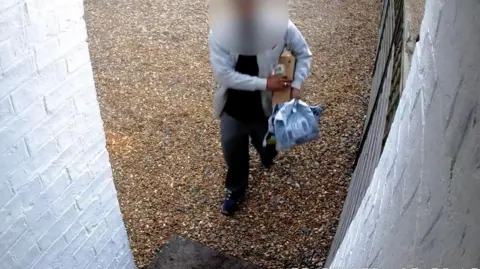 Toby Bennett A security camera image of a driver delivering parcels to a house. They are wearing dark grey trousers and top and a light grey jacket and their face has been blurred.