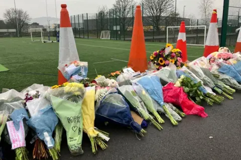 Dozens of bouquets of flowers are laid in front of the sports pitch. Orange traffic cones separate the flowers on the pavement and the field.